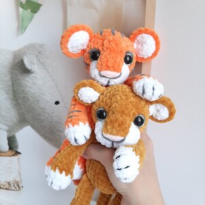 May include: Two crocheted tiger plushies, one orange and white and one brown and white, are stacked on top of each other. The orange tiger is on top and has a white face with black eyes and a pink nose. The brown tiger is on the bottom and has a white face with black eyes and a pink nose.