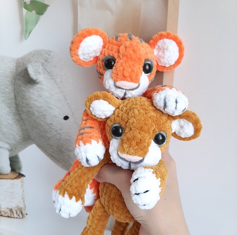 May include: Two crocheted tiger plushies, one orange and white and one brown and white, are stacked on top of each other. The orange tiger is on top and has a white face with black eyes and a pink nose. The brown tiger is on the bottom and has a white face with black eyes and a pink nose.