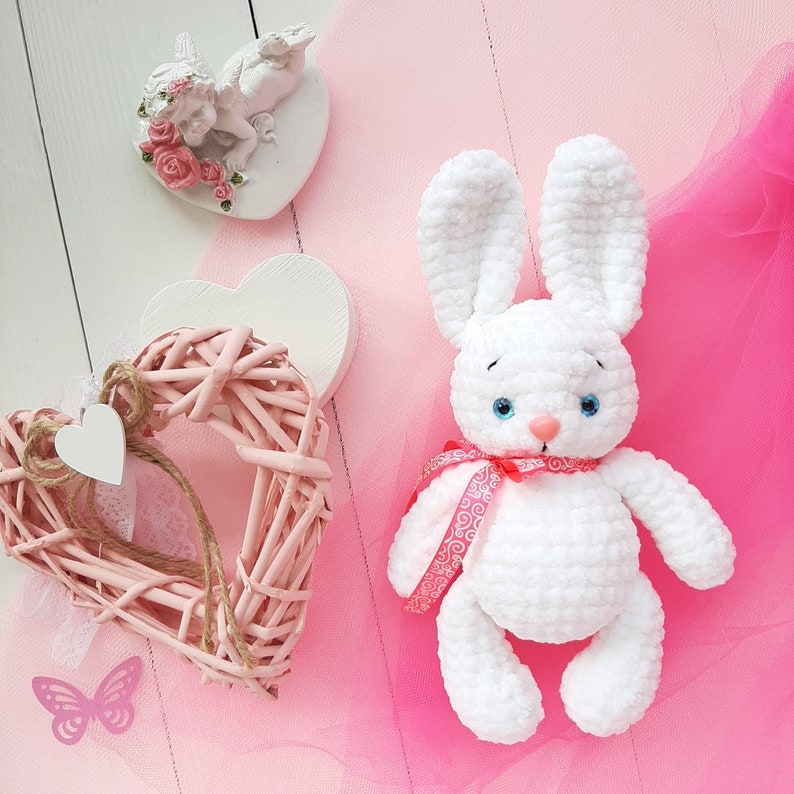 May include: White crocheted bunny rabbit toy with a pink and white ribbon. The bunny is sitting on a pink tulle fabric with a white wooden heart and a white angel figurine in the background.