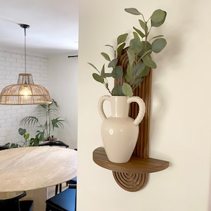 May include: A white ceramic vase with two handles sits on a wooden shelf with a curved design. The shelf is mounted on a white wall. A potted plant with green leaves is in the vase.
