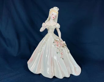 May include: A porcelain figurine of a bride wearing a white gown with lace detailing and a veil. She is holding a bouquet of pink flowers.