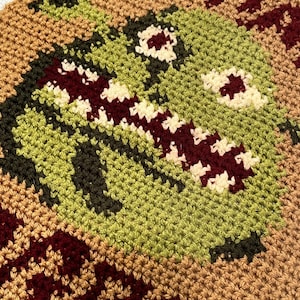 May include: Crocheted wall hanging with a green, brown, and red pixelated design of a monster face.