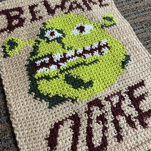 May include: A crocheted wall hanging featuring a green ogre with a toothy grin and the words "Beware Ogre" in brown yarn.