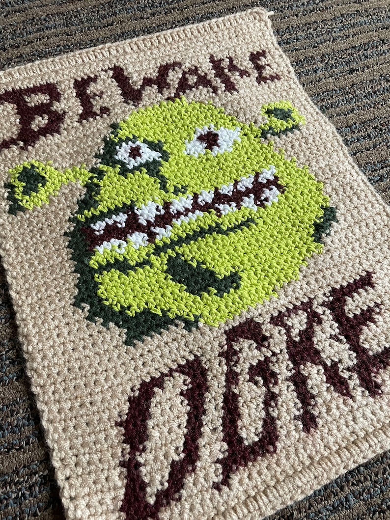 May include: A crocheted wall hanging featuring a green ogre with a toothy grin and the words "Beware Ogre" in brown yarn.