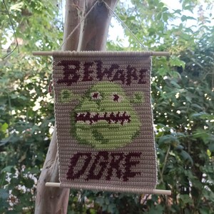 May include: A crocheted wall hanging with the words "Beware Ogre" in brown yarn. The ogre is green with a toothy grin and is surrounded by a beige background.