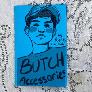 May include: A blue card featuring a hand-drawn illustration of a person wearing a cap. The text "BUTCH Accessories" is written in black, along with "by Ripley La Rue". The artwork is set against a blue background.
