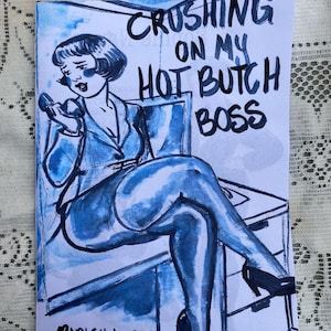 May include: A blue and white comic book illustration titled "Crushing on My Hot Butch Boss" by Ripley Larue. The artwork depicts a woman in a suit holding a phone, with a stylized, vintage aesthetic.