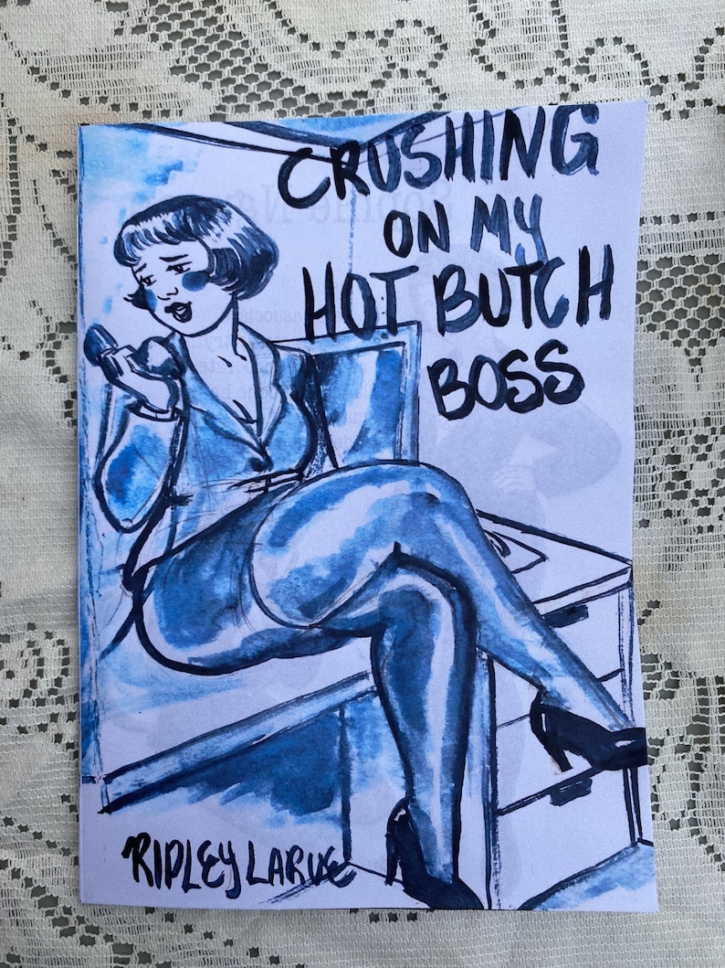 May include: A blue and white comic book illustration titled "Crushing on My Hot Butch Boss" by Ripley Larue. The artwork depicts a woman in a suit holding a phone, with a stylized, vintage aesthetic.