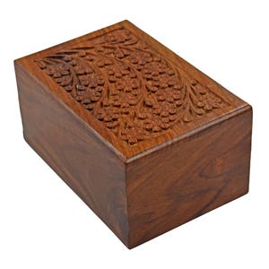 Puede incluir: Una urna de cremaci&oacute;n de madera con un dise&ntilde;o floral tallado. La urna est&aacute; hecha de madera marr&oacute;n oscura y tiene una forma rectangular.