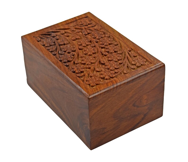 Puede incluir: Una urna de cremaci&oacute;n de madera con un dise&ntilde;o floral tallado. La urna est&aacute; hecha de madera marr&oacute;n oscura y tiene una forma rectangular.