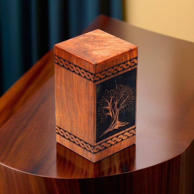 May include: A wooden box with a dark wood inlay featuring a tree of life design. The box is decorated with a Celtic knot border.