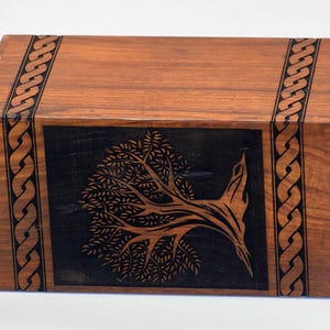 May include: A wooden box with a black inlay of a stylized tree design. The box has a Celtic knot border around the edges.