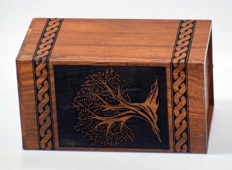 May include: A wooden box with a black inlay of a stylized tree design. The box has a Celtic knot border around the edges.
