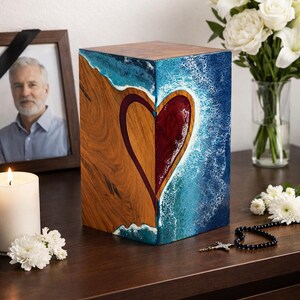 May include: A wooden cremation urn with a heart design. The urn is a cube shape, with a heart design in the center. The heart is divided into two halves, one side is wood grain, the other is a red hue. The background is blue and white.