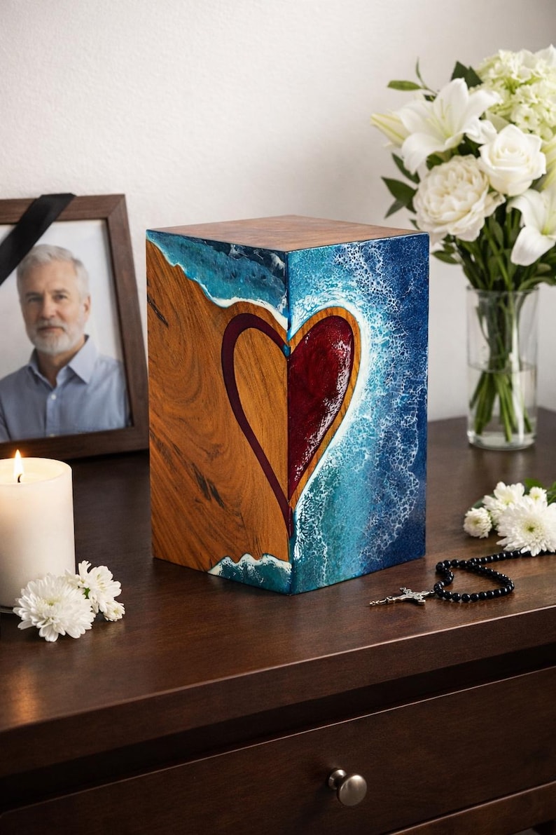 May include: A wooden cremation urn with a heart design. The urn is a cube shape, with a heart design in the center. The heart is divided into two halves, one side is wood grain, the other is a red hue. The background is blue and white.