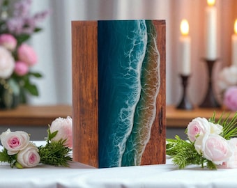 May include: A wooden box with a resin ocean design. The box has dark wood grain sides and a blue and white resin design resembling ocean waves and a sandy shore. The box is displayed on a white tablecloth with flowers.