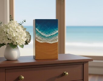 May include: A rectangular wooden art piece depicting a beach scene. The top portion shows deep blue ocean water transitioning to turquoise waves and white foam. The bottom half is a sandy beach. The artwork is displayed on a brown table.
