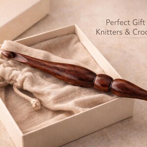 May include: A large, dark wood crochet hook rests inside a beige box with a matching drawstring bag. The hook has a smooth, polished finish and a rounded handle. Text in the image reads: "Perfect Gift for Knitters & Crocheters."