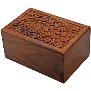 Puede incluir: Una urna de cremaci&oacute;n de madera con un dise&ntilde;o floral tallado. La urna est&aacute; hecha de madera marr&oacute;n oscura y tiene una forma rectangular.