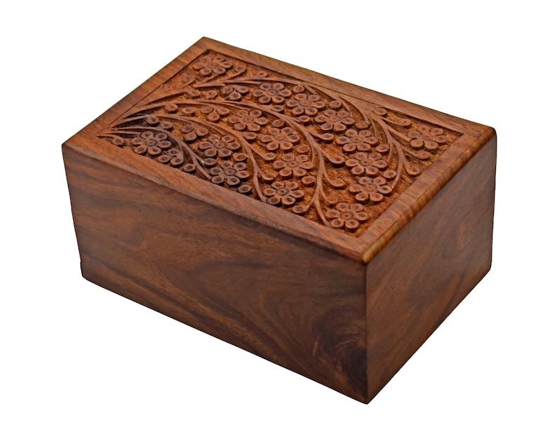 Puede incluir: Una urna de cremaci&oacute;n de madera con un dise&ntilde;o floral tallado. La urna est&aacute; hecha de madera marr&oacute;n oscura y tiene una forma rectangular.