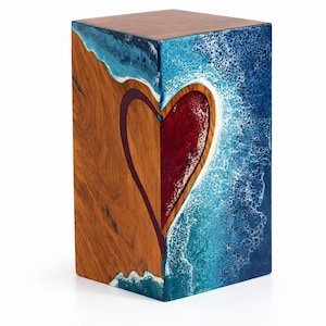 May include: A wooden cube with a heart design. One side features a heart shape in red, outlined in a dark color, set against a wood grain background. The other side depicts a blue ocean scene with white waves.