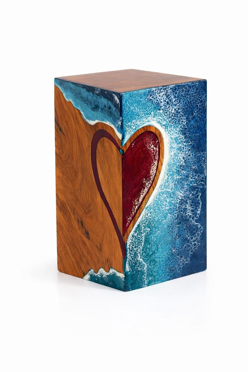 May include: A wooden cube with a heart design. One side features a heart shape in red, outlined in a dark color, set against a wood grain background. The other side depicts a blue ocean scene with white waves.