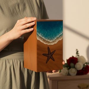 Pode incluir: Caixa de madeira com um design de cena de praia. A parte superior apresenta um oceano azul com ondas brancas, que se transforma numa praia arenosa com uma estrela do mar. A caixa é segurada por uma pessoa.