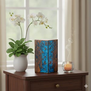 Może przedstawiać: Dekoracyjne drewniane pudełko z niebieskimi kwiatowymi wzorami i białym blatem stoi na ciemnobrązowym stole. Biała roślina orchidei w białej doniczce i zapalona świeca w szklanym świeczniku również znajdują się na stole.