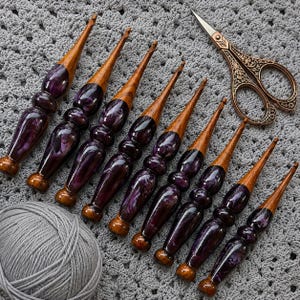 May include: A set of eight crochet hooks with polished wooden tips and ergonomic handles featuring a marbled deep purple and black resin design, accompanied by a pair of ornate, antique-style copper-colored metal embroidery scissors.