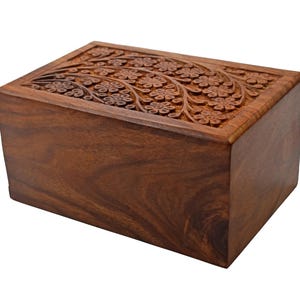 Puede incluir: Una urna de cremaci&oacute;n de madera con un dise&ntilde;o floral tallado. La urna est&aacute; hecha de madera marr&oacute;n oscura y tiene una forma rectangular.