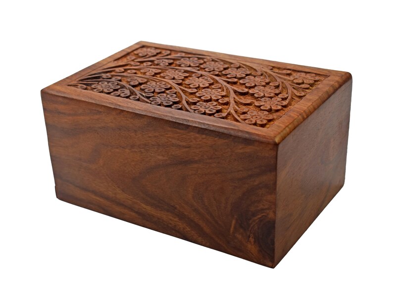 Puede incluir: Una urna de cremaci&oacute;n de madera con un dise&ntilde;o floral tallado. La urna est&aacute; hecha de madera marr&oacute;n oscura y tiene una forma rectangular.