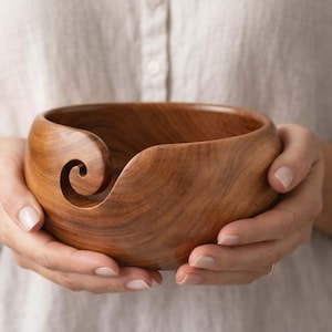 Puede incluir: Un cuenco de hilo de madera hecho a mano con un corte en espiral para el hilo. El cuenco es de madera de tonos cálidos y está sostenido por dos manos. Está diseñado para evitar que el hilo se enrolle mientras se teje o se hace ganchillo.