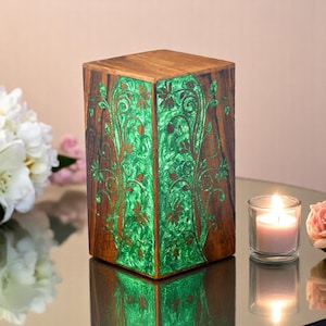 Könnte beinhalten: Eine Holzkiste mit einem grünen und braunen floralen Design. Die Kiste ist aus Holz und hat eine quadratische Form. Die Kiste steht auf einem Tisch mit einer Kerze im Hintergrund.