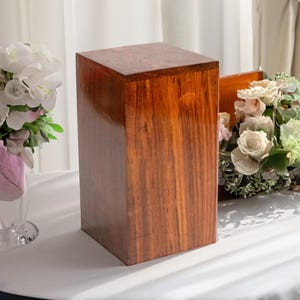 Puede incluir: Una caja de madera con un acabado marrón oscuro. La caja está sentada sobre un mantel blanco y está rodeada de flores.