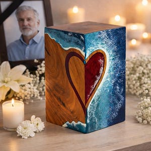 May include: A wooden urn with a heart design. The heart is divided, with one side featuring natural wood grain and the other side a deep red. The edges are detailed with blue resin, resembling ocean waves. A memorial photo and candles are in the background.