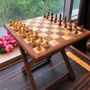 May include: A wooden chess set with a folding table. The chessboard is made of light and dark wood squares, with chess pieces set up for a game. The table has a dark brown finish and is supported by folding legs.