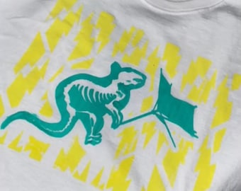 May include: White t-shirt featuring a green dinosaur skeleton graphic and yellow lightning bolt designs.