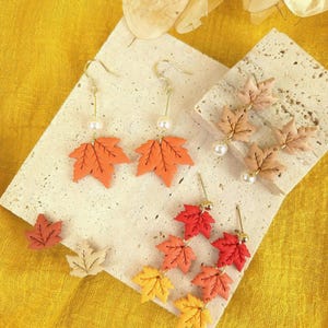 Peut inclure: Une collection de boucles d'oreilles et de clous en forme de feuille d'érable dans diverses couleurs, notamment orange, rouge, jaune et beige. L'ensemble comprend des boucles d'oreilles pendantes avec des perles et des crochets dorés.