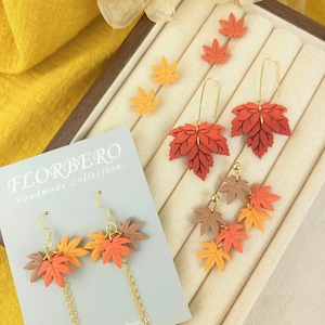 May include: Autumnal-themed earrings featuring maple leaf designs in orange, red, and brown. Some earrings have gold chains. The image also shows leaf-shaped stud earrings. The text "FLORBERO - Handmade Collection-" is visible.