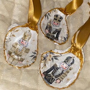 May include: Three oyster shell Christmas ornaments featuring nutcracker soldier designs. Each shell is painted with a different nutcracker, accented with gold trim and a gold ribbon for hanging. The ornaments have a festive, handcrafted appearance.