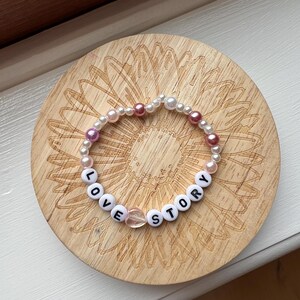 May include: A beaded bracelet with the words "LOVE STORY" spelled out in white letter beads. The bracelet also features white and pink beads, and a clear heart-shaped bead. The bracelet is resting on a wooden coaster.