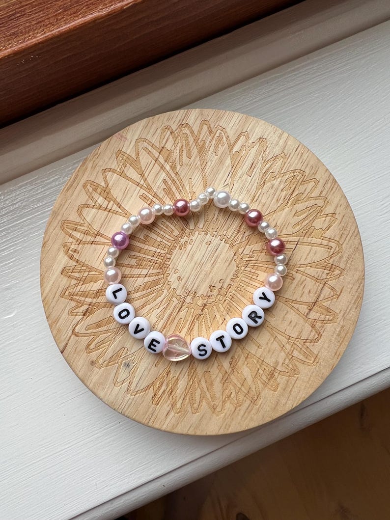 May include: A beaded bracelet with the words "LOVE STORY" spelled out in white letter beads. The bracelet also features white and pink beads, and a clear heart-shaped bead. The bracelet is resting on a wooden coaster.
