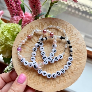May include: Three beaded bracelets with white letter beads spelling "WIFE", "BRIDES", and "WEDDING" on a wooden coaster. The bracelets also feature pearls and other beads in pink, black, and clear colors. Pink flowers are in the background.