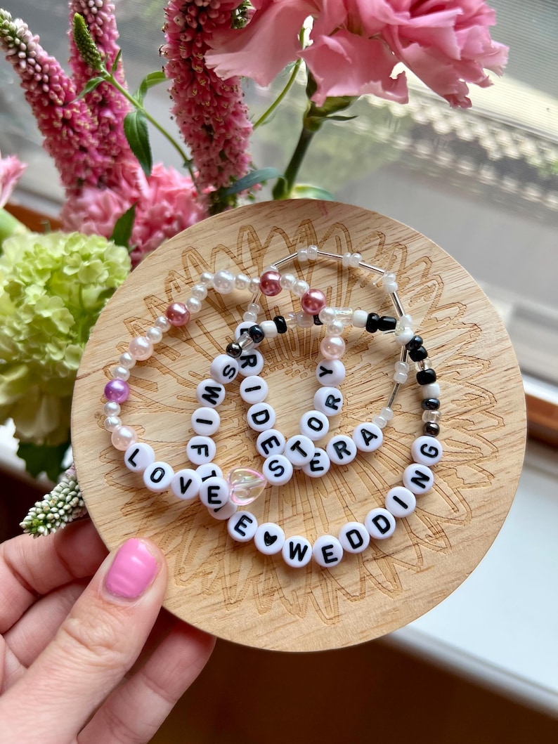May include: Three beaded bracelets with white letter beads spelling "WIFE", "BRIDES", and "WEDDING" on a wooden coaster. The bracelets also feature pearls and other beads in pink, black, and clear colors. Pink flowers are in the background.