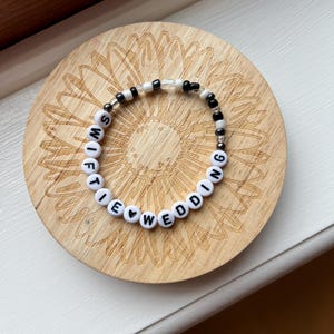 May include: A beaded bracelet with white letter beads spelling "SWIFTIE WEDDING" and black and clear beads. The bracelet is on a wooden coaster with a sunflower design.