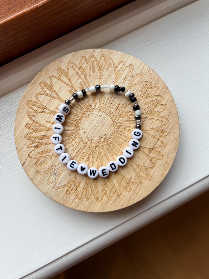 May include: A beaded bracelet with white letter beads spelling "SWIFTIE WEDDING" and black and clear beads. The bracelet is on a wooden coaster with a sunflower design.