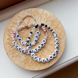 May include: Three beaded bracelets with white letter beads spelling "LOVE STORY", "SWIFTIE", and "ERA". The bracelets are on a wooden coaster with a floral design.