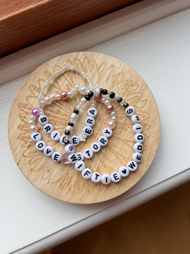 May include: Three beaded bracelets with white letter beads spelling "LOVE STORY", "SWIFTIE", and "ERA". The bracelets are on a wooden coaster with a floral design.