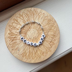 May include: A beaded bracelet spelling "BRIDE" with white letter beads and a star bead, strung on a clear elastic cord. The bracelet rests on a round wooden coaster with a sunflower design.