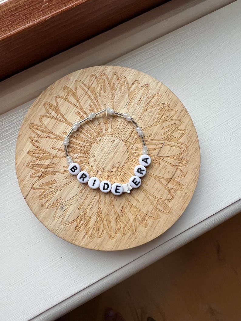 May include: A beaded bracelet spelling "BRIDE" with white letter beads and a star bead, strung on a clear elastic cord. The bracelet rests on a round wooden coaster with a sunflower design.